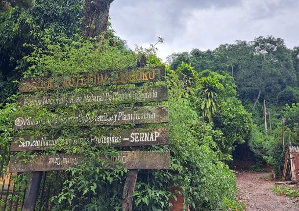 Weathered wooden sign for a Bolivian protected area, listing the National Service of Protected Areas (SERNAP), at the edge of a forest track